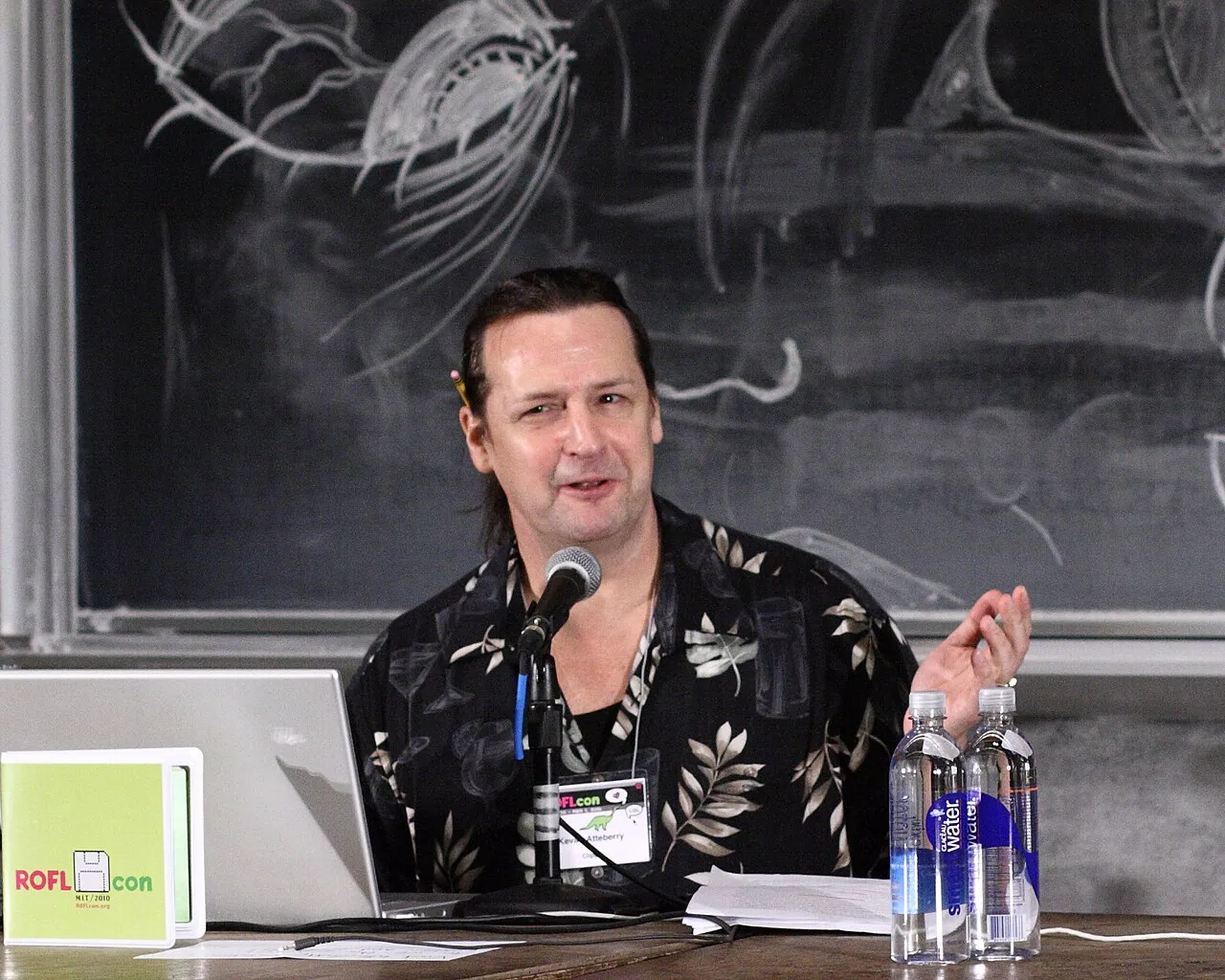 Illustrator Kevan Atteberry, who drew Clippy, speaking into a microphone at a table during the ROFLCon II internet-culture conference in 2010. He wears a black shirt with a leaf print, has a laptop and two water bottles in front of him, and a blackboard covered in chalk drawings behind him.