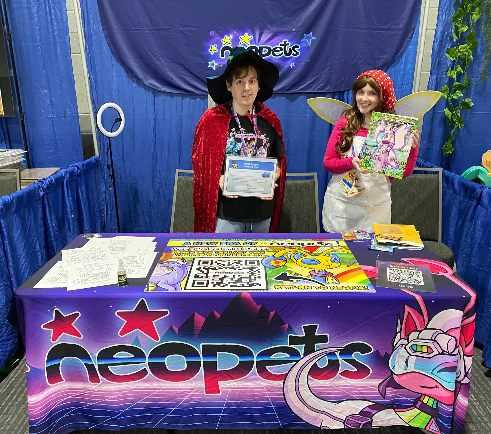 Two costumed Neopets representatives stand behind the purple Neopets booth at RetroGameCon 2023 with a tablet, coloring sheets, and scavenger-hunt signs.