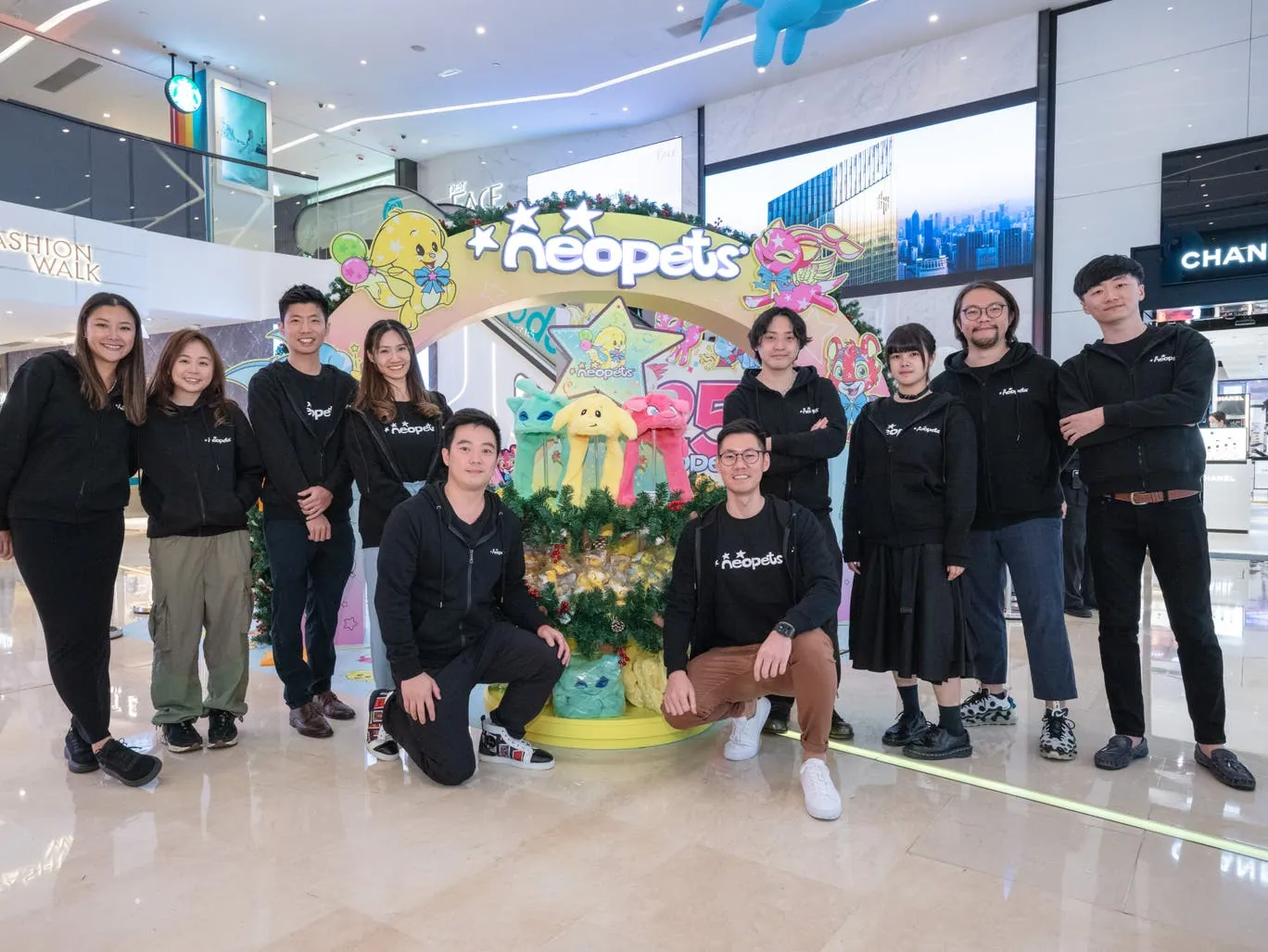 Members of the Neopets development team posing together beneath a colorful Neopets display in Hong Kong.